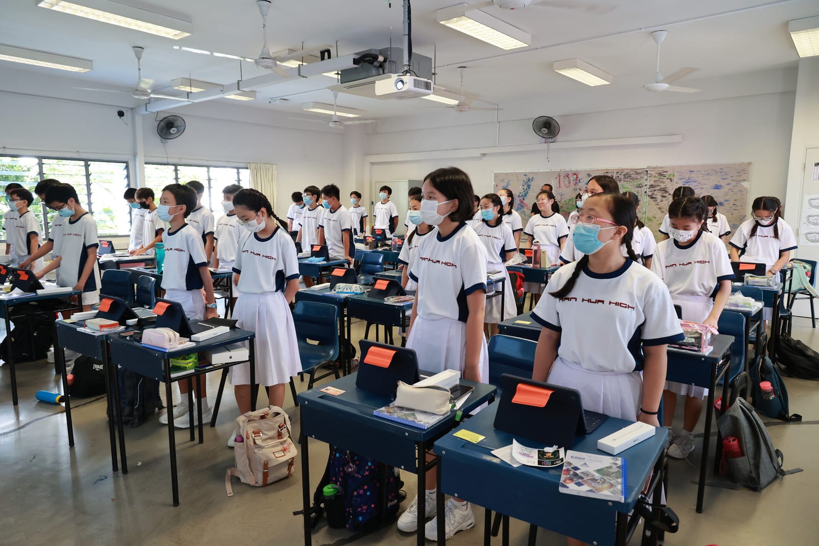Students in white uniforms and face masks stand at their desks in a bright classroom with a ceiling projector.